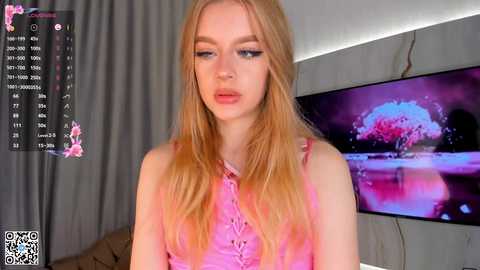 Media: Video of a young, fair-skinned blonde woman with long, wavy hair wearing a pink top, standing in front of a TV screen displaying a pink flower.