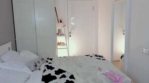 Media: Video of a minimalist, white-walled bedroom with a bed covered in a black-and-white cowprint duvet. A white wardrobe and a small shelf with toiletries are visible. The room has a clean, modern aesthetic.