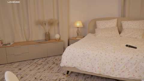 Media: Video of a cozy bedroom with beige walls, a plush bed, white floral-patterned bedding, a wooden nightstand with a lamp, and a small table with a white vase and decorative items.