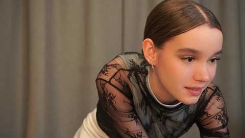 Media: Video of a young woman with fair skin and straight brown hair, wearing a sheer black lace top over a white shirt, leaning forward with a contemplative expression.