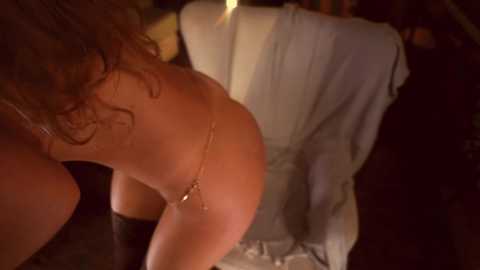 Media: Video of a nude woman with wet, curly hair, wearing a gold chain thong, bending over in a dimly lit room. A white garment hangs on a hanger in the background.