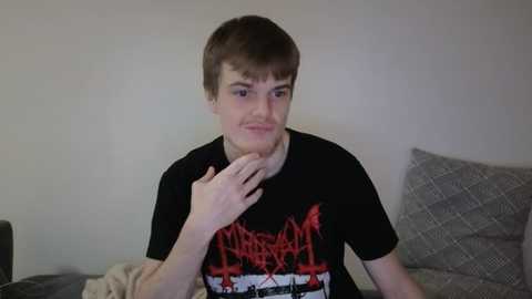 Media: Video of a young Caucasian man with short brown hair, wearing a black Metallica T-shirt, sitting on a gray couch against a plain white wall. He has a thoughtful expression, hand near his chin.