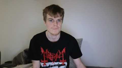 Media: Video of a young, fair-skinned man with short brown hair wearing a black T-shirt with a red and white graphic design. He sits on a bed in a plain, beige room.