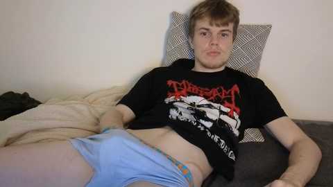 Media: Video of a young Caucasian man with short brown hair, wearing a black Metallica t-shirt and blue boxers, reclining on a bed with a beige blanket and grey pillow.