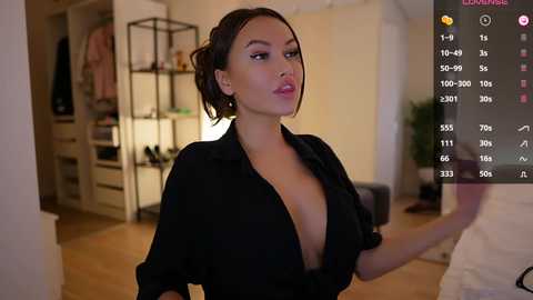 Media: Video of an Asian woman with medium skin tone and short black hair, wearing a black, low-cut blouse, standing in a modern, minimalist bedroom with wooden floors and a white bed.