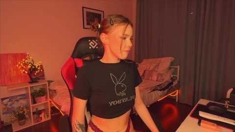 Media: Video of a blonde woman in a black Playboy Bunny crop top, sitting in a gaming chair in a cozy, dimly lit bedroom with a bed and shelves in the background.