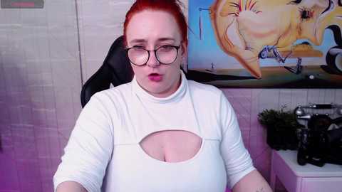 Media: Video of a plus-sized, red-haired woman with glasses, wearing a white, cut-out top, sitting in a chair in a studio with a wall painting of a nude woman and camera equipment.