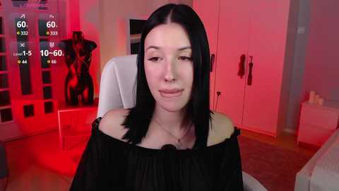 Media: A video of a fair-skinned woman with long black hair, wearing a black off-shoulder top, sitting in a modern room with red lighting and a mannequin in the background.