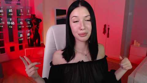 Media: Video of a young woman with long black hair, fair skin, and a slender physique, wearing a black off-shoulder top, sitting in a white chair. Background features a red-lit room with a wall display and a mannequin.