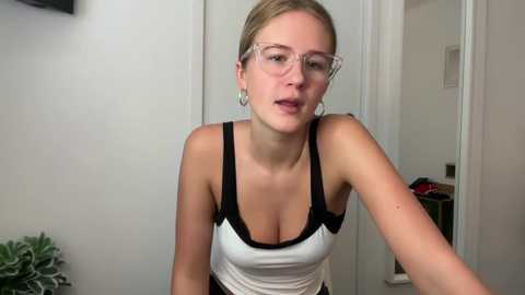 Media: Video of a blonde, fair-skinned woman with glasses, medium breasts, wearing a white tank top with black straps, in a simple indoor setting.