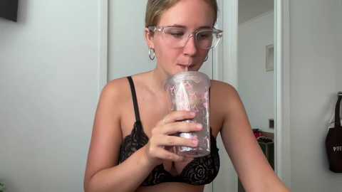 Media: Video of a fair-skinned, blonde woman with glasses, wearing a black lace bra, drinking from a transparent bottle in a modern, minimalist bathroom.