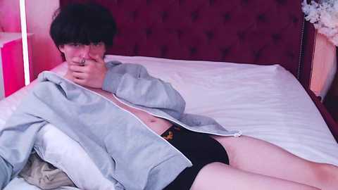 Media: Video of a young, fair-skinned man with short, dark hair, wearing a gray hoodie and black boxer shorts, lying on a bed with white sheets and a red headboard.