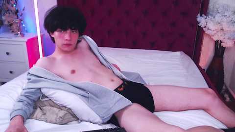 Media: Video of a young, slender, pale-skinned man with messy black hair, wearing an open grey shirt and black underwear, lying on a bed with white sheets and a red tufted headboard.