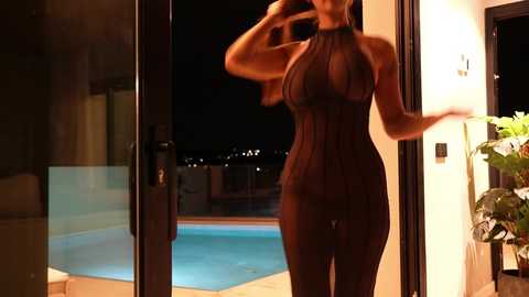 Media: Video of a slender woman with long, dark hair, wearing a sheer black halter top and matching pants, standing in a doorway, looking out at a nighttime pool with city lights in the background.