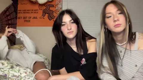 Media: Video of three young women with pale skin and long dark hair, one with a septum piercing, sitting on a bed, in casual attire, with a colorful wall poster behind them.