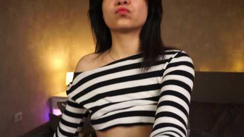 Media: Video of a young woman with straight black hair, wearing a black-and-white striped crop top, puckering her lips. Background shows a dimly lit bedroom with a bedside lamp and a gray headboard.