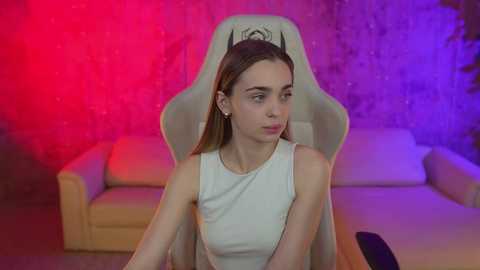 Media: Video of a young Caucasian woman with straight brown hair, wearing a white sleeveless top, sitting on a white gaming chair in a dimly lit room with red and purple lighting, surrounded by beige couches.