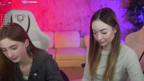 Media: Video of two young women in a cozy, colorful room. One with brown hair in a black jacket, the other with blonde hair in a beige top.