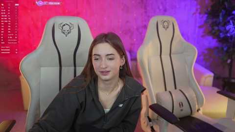 Media: Video of a young woman with fair skin, straight brown hair, wearing a black jacket and sitting in a white gaming chair with skull designs. Background shows a colorful, vibrant room with red and purple lighting, and a potted plant.