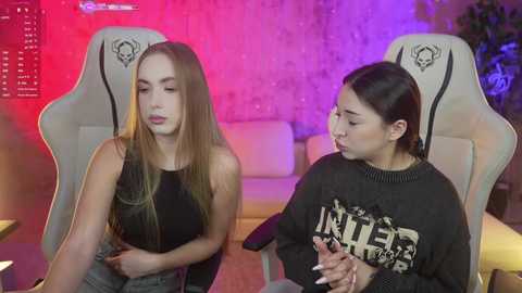 Media: Video of two young women in casual black clothing, seated on gaming chairs with a neon-lit background, one with long blonde hair and the other with dark hair, appearing tired and thoughtful.