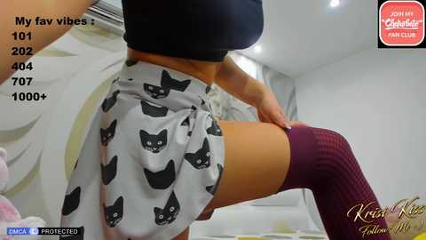 Media: Video of a woman in a black crop top, white skirt with black cat prints, and thigh-high maroon socks, posing indoors.