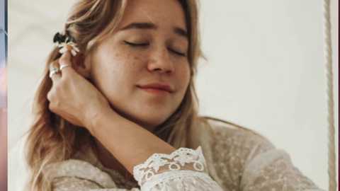 Media: Video of a young woman with long, wavy blonde hair, wearing a white lace blouse, resting her head on a man's shoulder, eyes closed, serene expression.