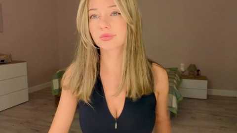 Media: Video of a blonde woman with fair skin, wearing a sleeveless black top with a zipper, standing in a minimalist bedroom with beige walls and a green and white checkered bedspread.