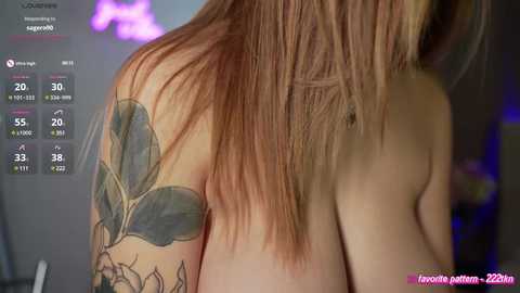 Media: Video of a topless woman with long, straight, light brown hair. She has a large, colorful tattoo of green leaves on her left shoulder. The background is blurred, showing a purple light and a pink text overlay.