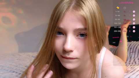 Media: A video of a young blonde girl with fair skin, blue eyes, and straight hair. She is lying on a bed, wearing a white tank top. The background includes a blurry, colorful pattern and digital interface elements.