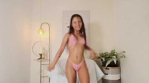 Media: Video of a smiling, slender, light-skinned woman with long dark hair, wearing pink lingerie, standing in a modern, minimalist room with white walls, a potted plant, and a white chair.