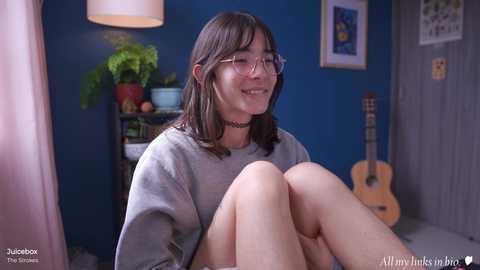 Media: Video of a young woman with straight brown hair and glasses, wearing a gray sweater and black choker, sitting on a bed in a blue-walled room with a guitar, plants, and framed art.