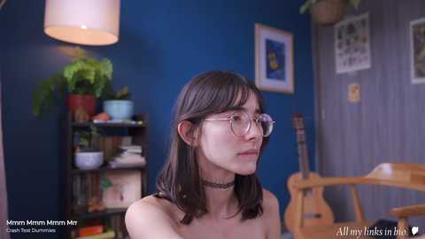 Media: Video of a young woman with shoulder-length dark hair, wearing glasses, a black choker, and a black top, in a colorful, cozy room with blue walls, potted plants, a guitar, and a wooden chair.