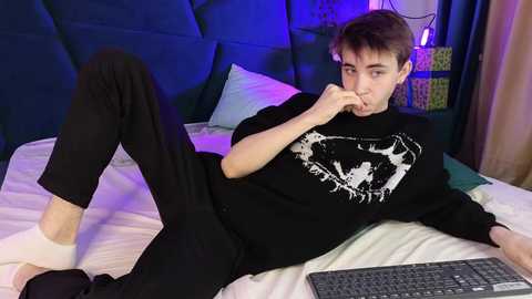 Media: Video of a young, thin man with short brown hair, lying on a bed, wearing black pants and a black graphic t-shirt, using a laptop, in a dimly lit bedroom with a blue-tinted headboard and colorful wall art.