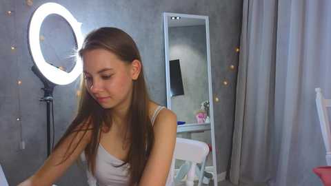 Media: Video of a young Caucasian woman with straight brown hair, wearing a gray tank top, standing in a modern bedroom with a large circular light, gray walls, a mirror, and a white chair.