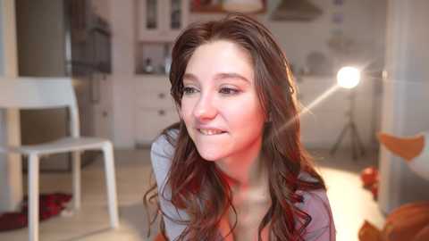 Media: Video of a young woman with long brown hair, wearing a gray top, smiling in a warmly lit room with a white chair, shelves, and a lamp.