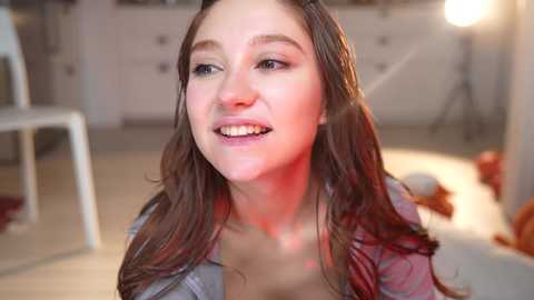 Media: Video of a young woman with long, wet brown hair, wearing a gray shirt, smiling, in a brightly lit room with a white chair and dresser in the background.