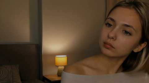 Media: Video of a young woman with fair skin and light brown hair, looking contemplative. She's in a dimly-lit room with a bedside table lamp and a headboard.