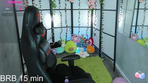 Media: A video captures a playful, cozy gaming room with a black gaming chair, colorful plush toys, green rug, and pink flowers, set against a soft pink and green backdrop.