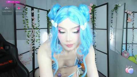 Media: Video of a light-skinned woman with blue hair styled in buns, wearing a floral bikini, and a flower headpiece, in a dimly-lit room with a pink and white theme, including a pink chair.