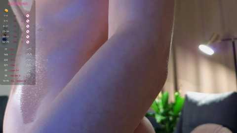 Media: A close-up video of a nude person with fair skin, highlighting their arm and torso, with a blurred background of a living room featuring a potted plant and a lamp.