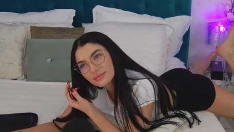 Media: Video of a slender, fair-skinned woman with long black hair, wearing glasses and a white top, lying on a bed with a teal headboard.