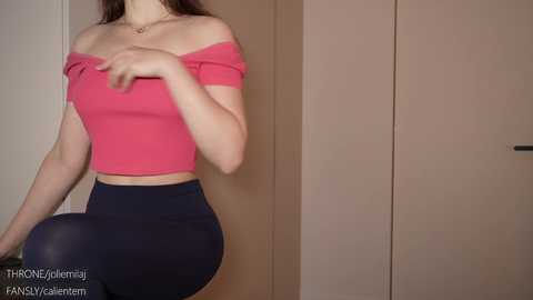 Media: Video of a fair-skinned woman with long brown hair, wearing a pink off-shoulder crop top and high-waisted navy blue pants, posing indoors against a beige wall and wooden wardrobe.