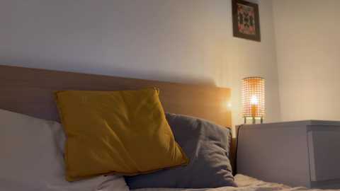 Media: Video of a minimalist bedroom featuring a wooden headboard with a mustard-yellow pillow and a blue pillow, lit by a modern, cylindrical lamp on the nightstand. A framed painting hangs on the wall.