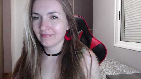 Media: A video of a fair-skinned, young woman with long, straight, light brown hair, wearing a black choker necklace, sitting in a gaming chair with red accents. She smiles softly.