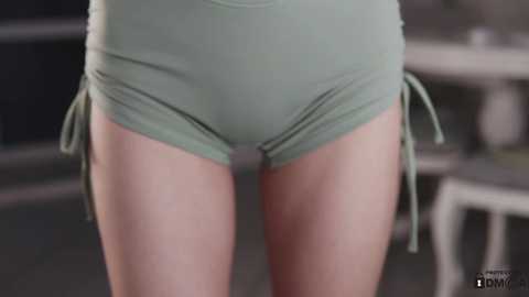 Media: Video of a woman's lower torso in mint green athletic shorts with side ties, standing indoors with blurred background.