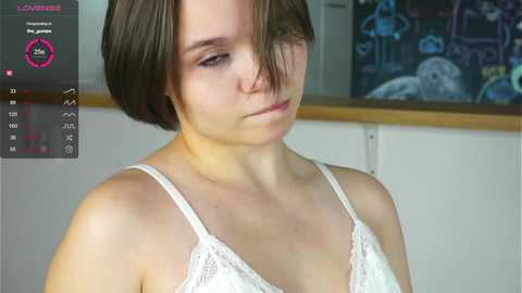 Media: Video of a young woman with short brown hair, wearing a white lace bra, looking down, in a room with a chalkboard and a digital display showing her vital signs.