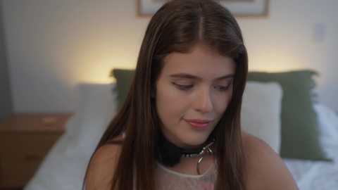 Media: Video of a young woman with long brown hair, wearing a black collar, sitting on a bed with white and green bedding, in a softly lit bedroom.