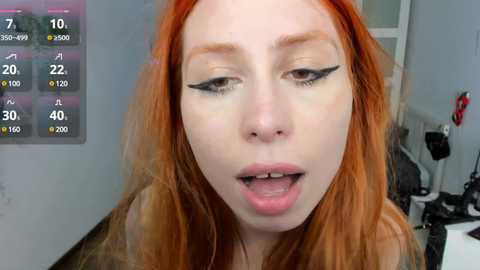 Media: Video of a fair-skinned, red-haired woman with heavy black winged eyeliner, slightly open mouth, in a studio setting with a white wall and a camera monitor.