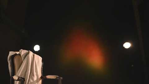 Media: Video of a dark, dimly lit outdoor scene at night, featuring a white towel draped over a wooden chair, with two bright spotlights creating a warm, orange glow in the background.