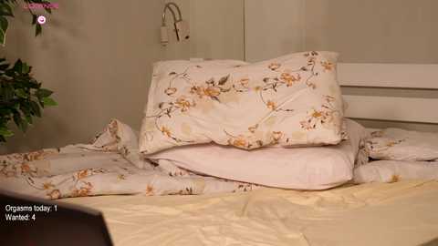 Media: A video shows a messy bed with a white duvet and pillows featuring orange floral patterns. The bed is in a beige room with a white headboard. A leafy green plant is on the left.